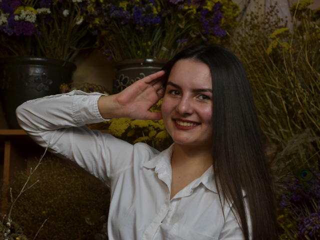 JasmineAsha with long dark hair, white shirt, smiling, hand near ear, surrounded by flowers, showing cute ass, tits barely visible under shirt, in a floral-filled setting.