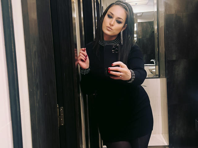 Elise Winfrey, con cabello largo y castaño, vestido negro y collar con tachuelas, apoyada en una puerta, sosteniendo un teléfono, mirando un espejo, usando medias negras, con una expresión seria..