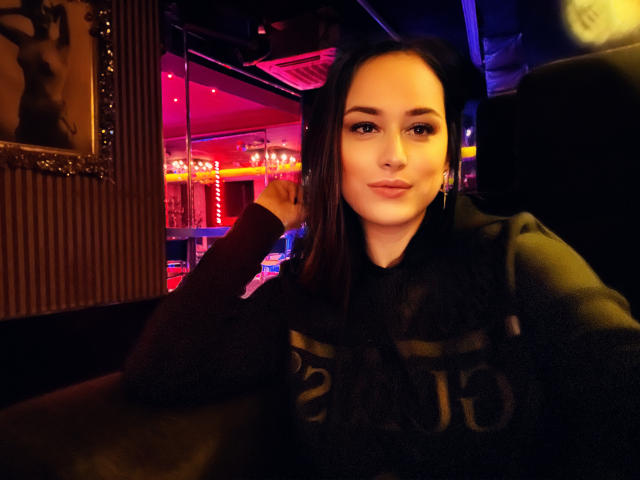 Elise Winfrey, con cabello castaño largo, vestida con una sudadera negra, tiene un rostro lindo, senos pequeños y una leve sonrisa, y está sentada en un bar tenuemente iluminado, con luces de neón..