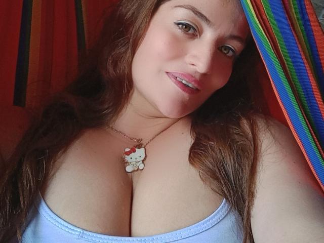 VikMe avec des cheveux bruns, un débardeur bleu, montrant de grandes seins, portant un collier Hello Kitty, souriant, regardant la caméra, rideau rayé arc-en-ciel sur la droite..