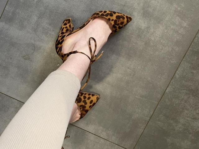 RozalinaPleasure, con un pie calzado en tacones puntiagudos de estampado de leopardo y finas tiras marrones, lleva una manga beige texturizada, sobre un suelo de baldosas grises. Su tobillo está ligeramente flexionado..