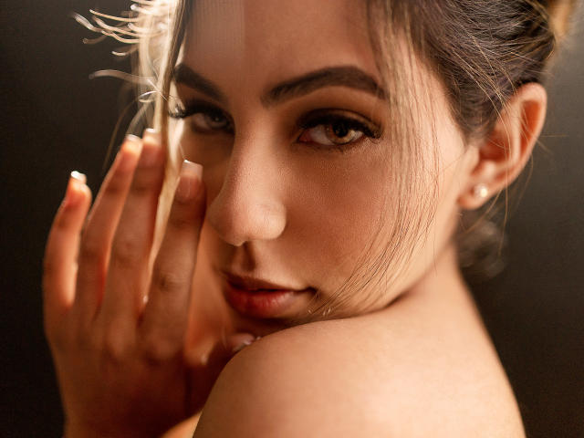Le visage de KataMillerr, ses yeux fixant de manière séduisante l'appareil photo, avec sa main partiellement recouvrant sa joue, mettant en valeur son épaule nue et laissant entrevoir ses seins..