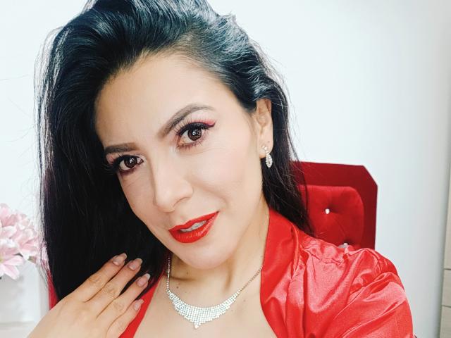 JoTaylor with red lipstick, shiny black hair, red satin shirt, sparkling necklace, and earrings, smiling, hand on chest, red chair, pink flowers in background, sexy, attractive.
