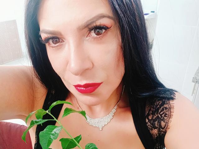 JoTaylor with dark hair, red lipstick, and a diamond necklace, holding a green plant, wearing black lace, brown eyes, and long eyelashes. Boobs visible in black lace top.