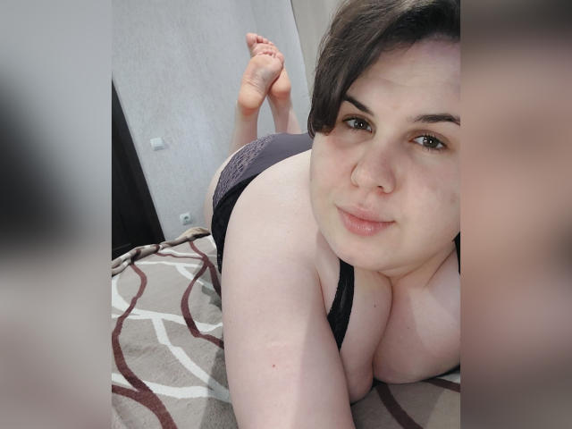 BeckyAndEllen, lying on her stomach, with her tits pressed against a bed, wearing black lace panties, her ass raised, short dark hair, nose ring, smiling at the camera.