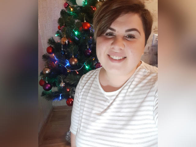 BeckyAndEllen with short brown hair, smiling, wearing a white striped shirt, standing in front of a decorated Christmas tree with colorful lights and ornaments.