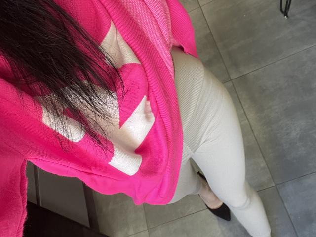 RozalinaPleasure's roze trui onthult haar blote borst, de witte, geribbelde broek omhelst haar strakke billen, ze draagt zwarte schoenen en heeft lang, donker haar. De vloer is grijs betegeld, en de witte letters op de roze trui zijn zichtbaar..
