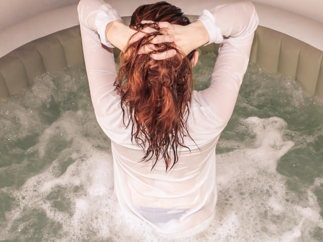 BKLove in una camicia bianca bagnata, con le mani tra i capelli castani, seduta in una vasca idromassaggio piena di acqua bollente, con l'acqua che le copre i glutei e la biancheria intima bianca. Scatto dall'alto..