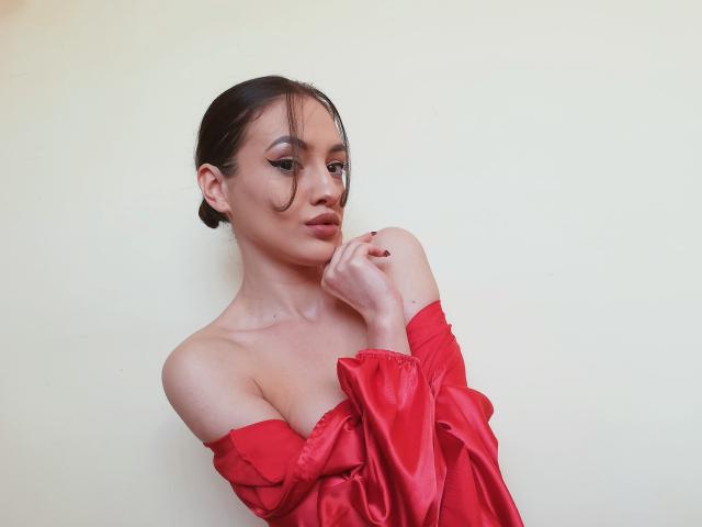 Vievienne Clemente, avec ses cheveux foncés attachés en chignon, portant un haut rouge à une épaule, les mains près de son visage, un maquillage discret, regardant légèrement sur le côté, un sourire doux, peau claire..