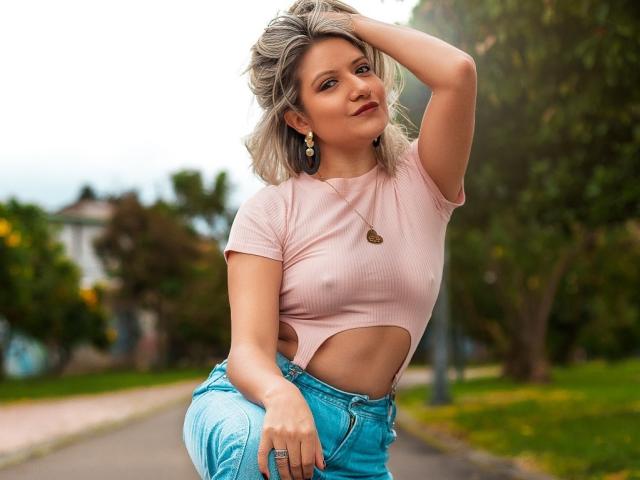 ValeriaHunter69 with blonde hair, pink crop top showing her tits, and blue jeans, hand in hair, smiling, standing on a sidewalk, green background, gold earrings, and a necklace.