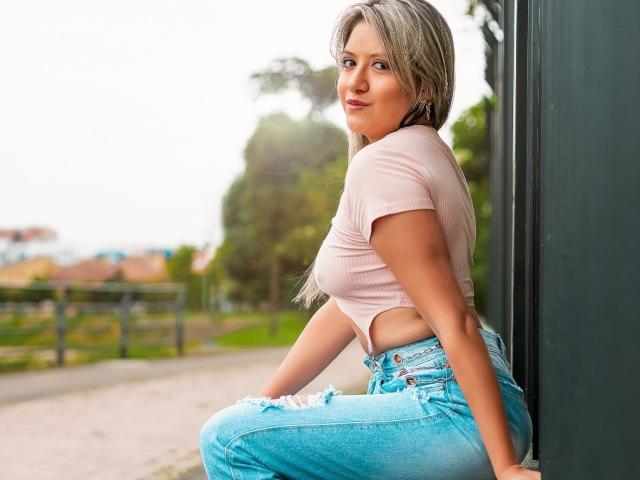 ValeriaHunter69 met blond haar, een roze geribbelde crop top die haar borst laat zien, en blauwe versleten jeans, zittend tegen een donkere muur, kijkt terug met een lichte glimlach..