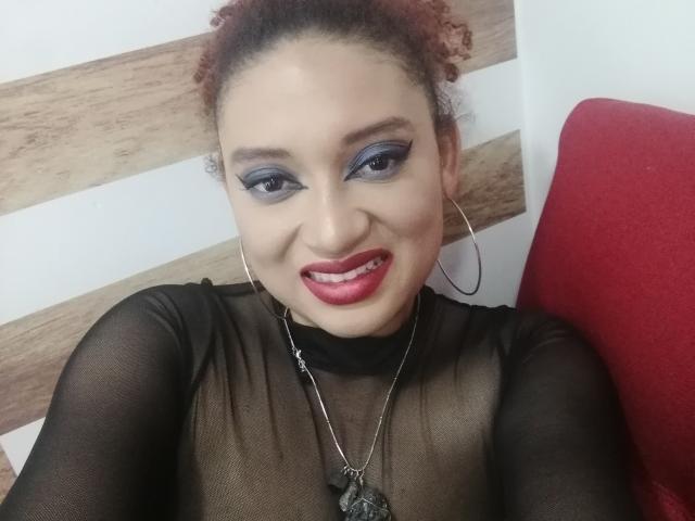 VioletaSwitch has curly red hair, blue eyeshadow, red lipstick, black sheer top, silver necklace, and hoop earrings, smiling at the camera with red chair on the right.