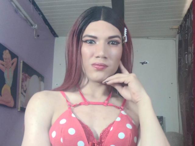 LunaGransTs con capelli rosso-marrone, indossa un reggiseno rosa a pois con pizzo rosso, si tocca il mento, guarda la fotocamera, mostrando il seno. Trucco leggero, rossetto rosa..