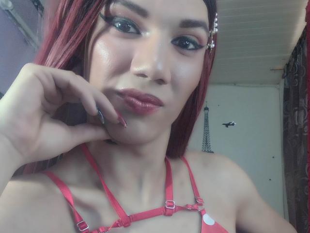 LunaGransTs with red hair, dark eyeliner, and pink lipstick, wearing a red harness, has a hand near her face, showing off her shiny skin and pink lips.