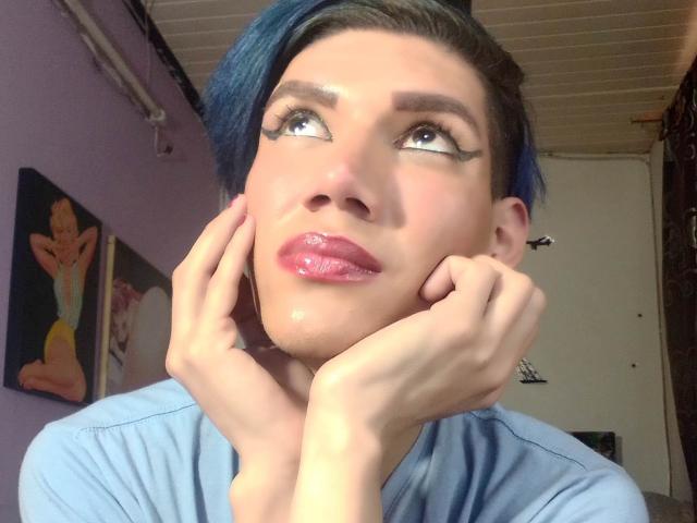 LunaGransTs with blue hair, glossy pink lips, and thick black eyeliner, looking up with hands on cheeks, wearing a light blue shirt, in a well-lit room.