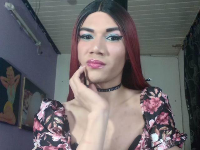 LunaGransTs with red hair, blue eye makeup, pink lipstick, floral dress, black choker, hand on cheek, medium tits, light skin, ceiling above, white wall behind.