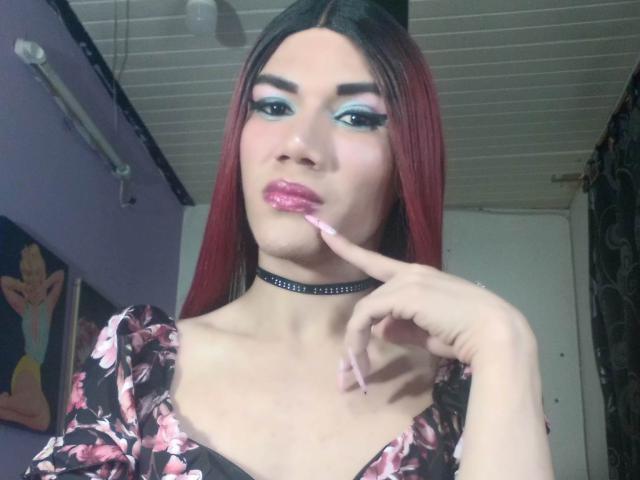 LunaGransTs with red hair, blue eyeshadow, pink lipstick, and floral dress, sucking her finger, wearing a black choker, in a room with white ceiling and purple walls.