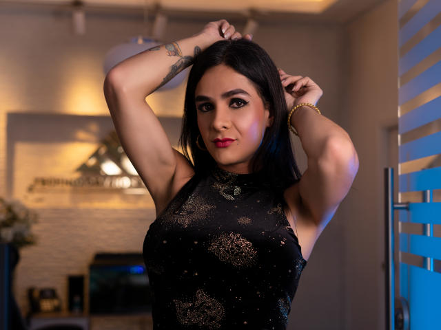 KristalWels com os braços levantados, vestindo um vestido preto sem mangas com padrões dourados, batom vermelho e um bracelete, em pé em um quarto com iluminação azul à direita..