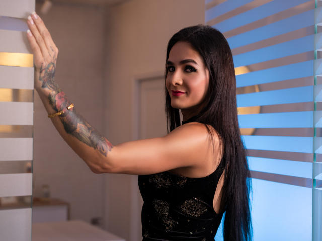 KristalWels, com cabelos pretos longos, vestido de renda preta, braço tatuado e batom vermelho, sorrindo para a câmera, em pé em frente a uma parede listrada de branco e azul. Sem seios aparentes, área genital não visível..