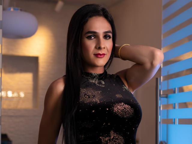 KristalWels with long black hair, wearing a black dress with gold patterns, red lipstick, and a gold bracelet, hand behind head, looking at camera, tits visible, medium breast size.