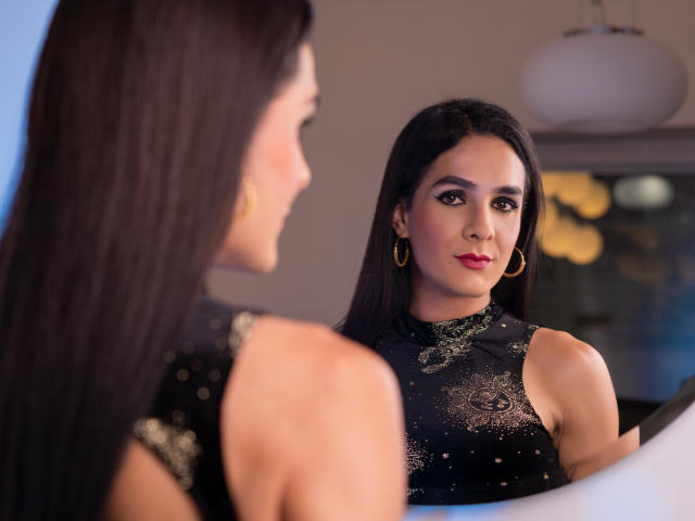 KristalWels with long black hair, wearing a black lace dress, gold hoop earrings, and red lipstick, stands in front of a mirror, gazing at her reflection.