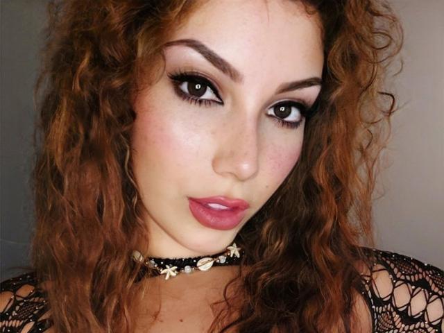 SexyRealWoman69, con i capelli castano ricci, indossa un collare di pizzo nero, ha il rossetto rosa, l'eyeliner scuro e un sorriso carino, guardando direttamente la fotocamera. Il seno è visibile attraverso il pizzo..