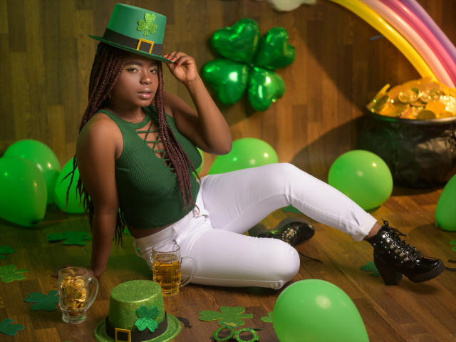 LindaBird com pele escura, tranças, chapéu verde, cropped verde, calças brancas, botas pretas, sentada em um chão de madeira, segurando uma cerveja, cercada por balões verdes e trevos..