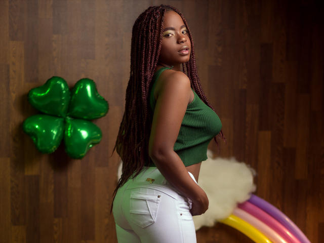 LindaBird with dark skin, long red braids, green sleeveless top, white jeans, looking back, showing her ass, standing in front of a green shamrock and rainbow.