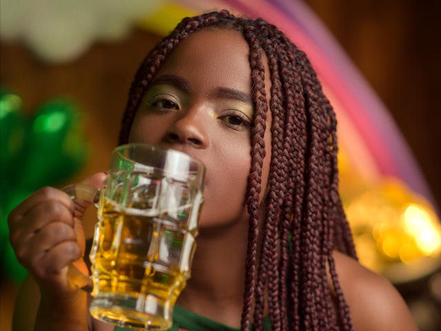 LindaBird, con i capelli castano intrecciati, beve da un bicchiere di birra dorata, con l'ombretto giallo-verde e la maglia verde ben visibili. Ha uno sguardo rivolto in avanti e un'espressione rilassata..