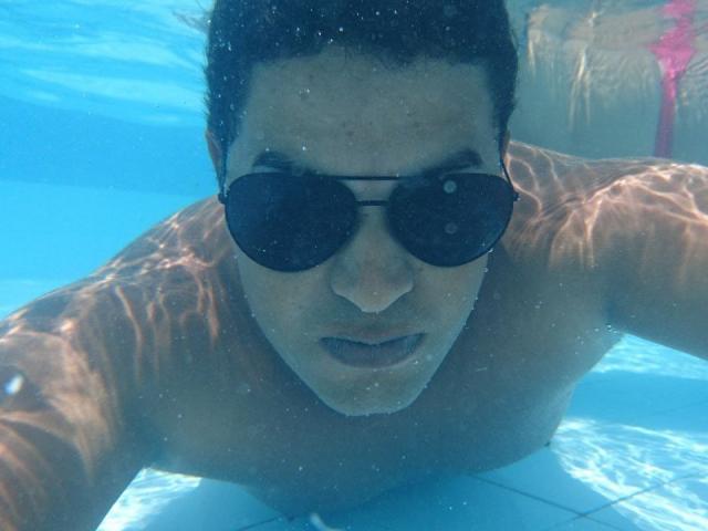 Unterwasseraufnahme von MrBlackHot mit dunklen Sonnenbrillen, brauner Haut und kurzen schwarzen Haaren, der flach liegt und einen ernsten Gesichtsausdruck hat, wobei Brust und Gesicht im klaren blauen Wasser liegen..