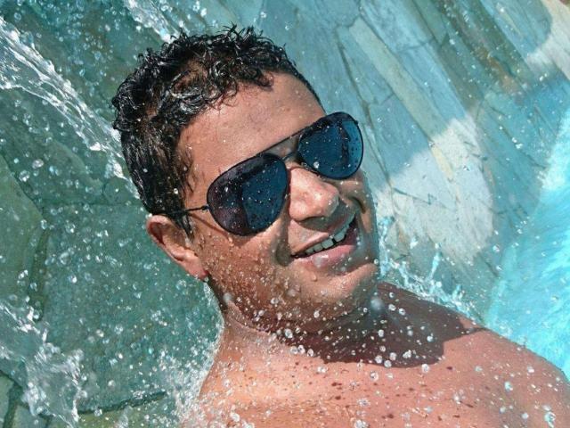 MrBlackHot with wet, curly dark hair, wearing black sunglasses, smiling underwater, water droplets splashing his shirtless, tanned, muscular upper body.