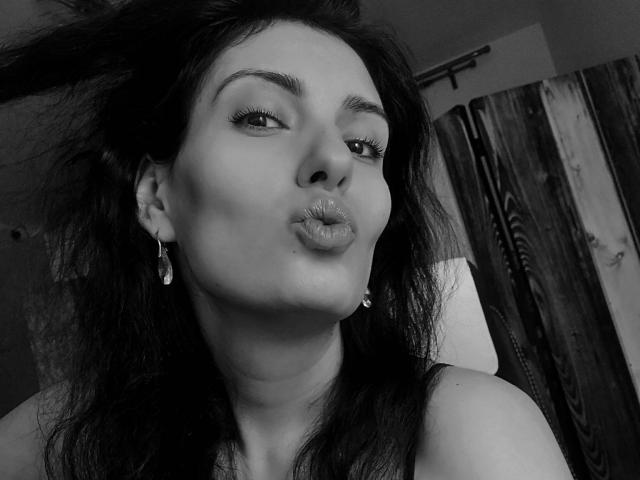 Black-and-white photo of AddictedToUTS with dark, messy hair, pouty lips, and small earrings, slightly tilting head, wearing a sleeveless top, looking at camera with a playful expression.