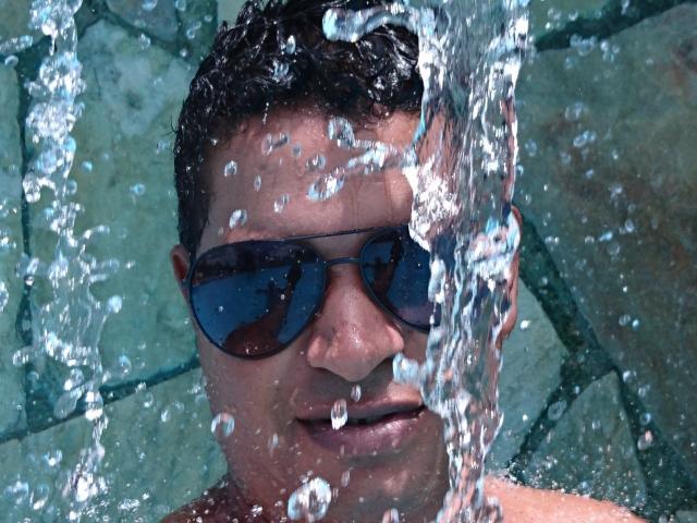 MrBlackHot, com cabelo curto e escuro, usando óculos de sol escuros, tem água espirrando em seu rosto. Ele está sem camisa, com gotas cobrindo o tronco superior e uma expressão neutra..