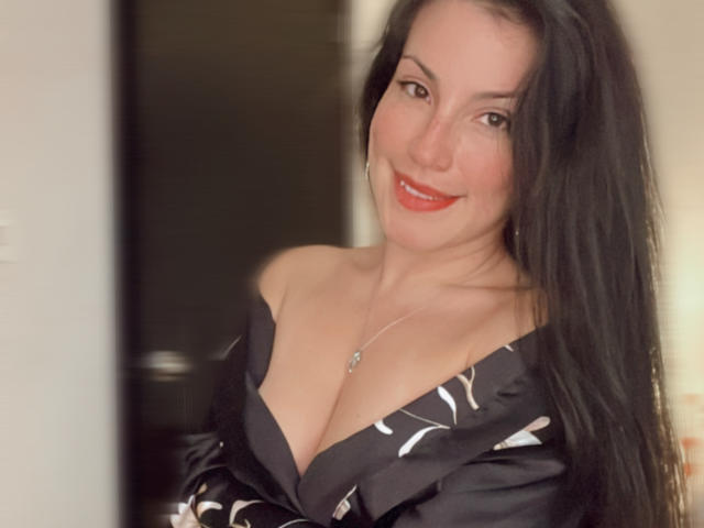 Liliysweet, com longos cabelos pretos, sorrindo, vestindo uma blusa preta com decote ombro a ombro e estampas brancas, mostrando seus seios, batom vermelho e um colar de prata, em uma foto com iluminação suave..