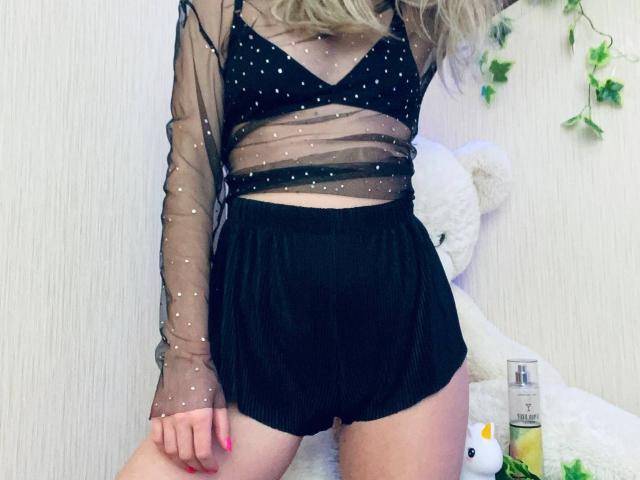 AlicePrings with blonde hair, wearing a sheer black polka dot top and black high-waisted shorts, exposing her tits and ass, against a white wall with a teddy bear.