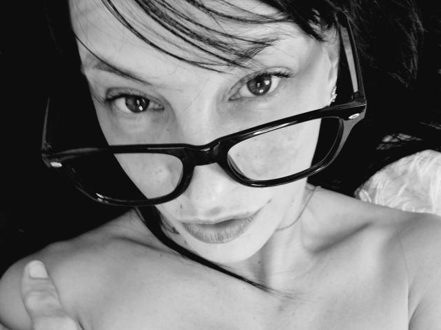Autoportrait en noir et blanc de CassandraMichelli, portant des lunettes de soleil, avec des taches de rousseur et un léger sourire, montrant ses épaules dénudées et levant le pouce. Elle a les cheveux foncés et une bouche discrète..