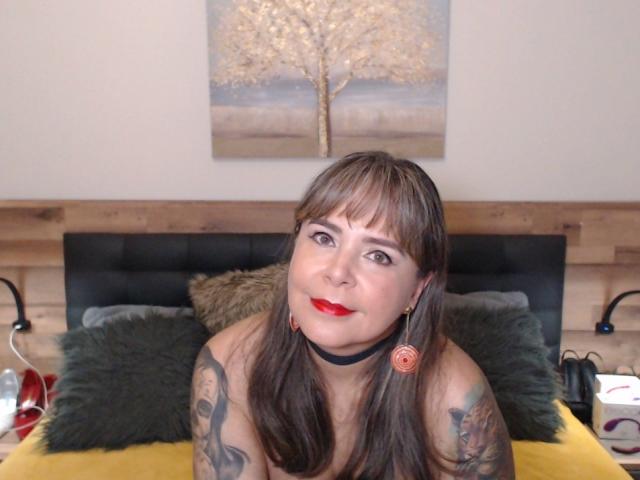 EvaLuv avec du rouge à lèvres, des tatouages sur les bras, des cheveux bruns avec une frange, un collier noir, des boucles d'oreilles, nue, sur un lit jaune, avec des oreillers verts, une tête de lit noire, un mur en bois, souriante..