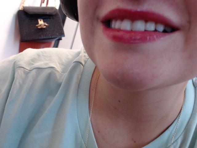 O rosto de LuxEsa, com batom vermelho, dentes brancos e um colar de ouro, vestindo uma camisa verde clara, com uma bolsa preta e fones de ouvido ao fundo..