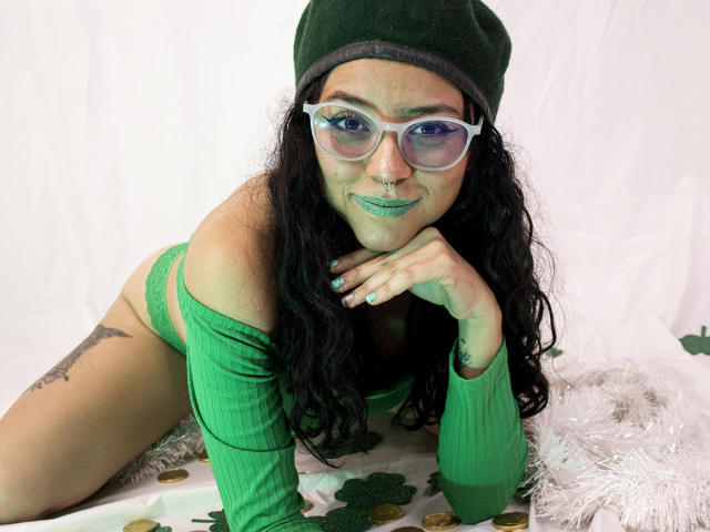 SamyBrook, com batom verde, óculos e gorro, deitada em um tecido verde e branco, vestindo uma blusa verde com um ombro de fora, mostrando os seios, calcinha verde e tatuagens, sorrindo com a mão no queixo..