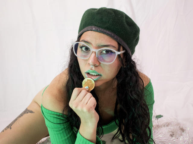 SamyBrook, com batom verde, óculos e um gorro verde, segurando um pirulito, tem cabelos longos e cacheados pretos e uma tatuagem no braço direito, vestindo uma blusa verde com um ombro de fora..