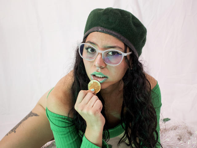 SamyBrook com gorro verde, óculos, batom verde, segurando um pirulito, blusa verde, cabelo preto ondulado, piercing no nariz, tatuagem no braço direito, fundo branco. Seios parcialmente visíveis..