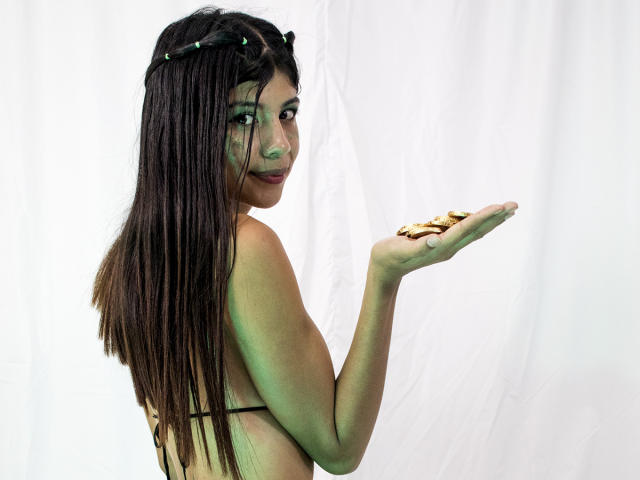 SamyNicols, con cabello castaño largo y trenzado, maquillaje de ojos verde, sosteniendo un puñado de monedas de oro, mirando hacia atrás con una ligera sonrisa, vistiendo un top de bikini negro de tiras..
