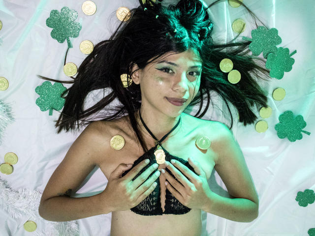 SamyNicols lying on a white sheet, wearing black lace bra, covering her tits with her hands, green light on her face, coins and green shamroks around her.