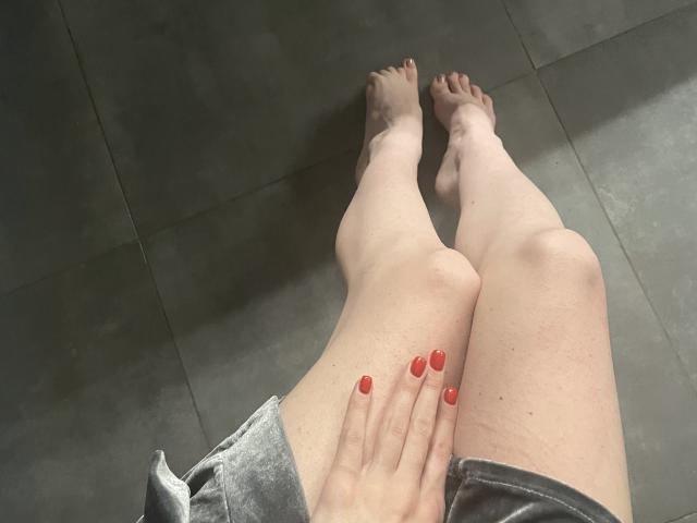 RozalinaPleasure's Beine mit heller Haut, rot lackierten Nägeln und grauen Shorts, die Füße auf einem dunklen Fliesenboden, die Zehen leicht gespreizt. Ihre rechte Hand ruht auf ihrem linken Oberschenkel..
