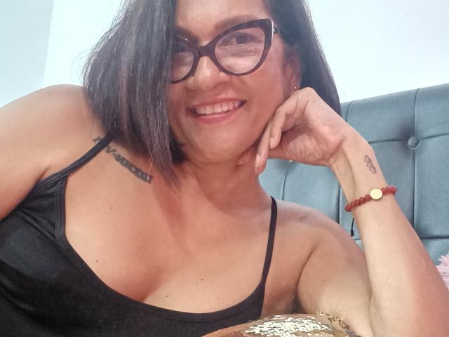 MatureJennyX with short black hair, glasses, and a black tank top, showing her tits and tattoos, smiling, resting her chin on her hand, wearing a brown bracelet.