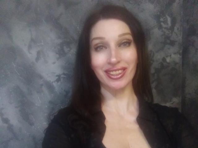 Jillian Pattie mit dunkelbraunen Haaren, blauen Augen und einem Lächeln, trägt ein schwarzes Oberteil, das ihre Brüste zeigt, vor einem strukturierten, grauen Hintergrund..