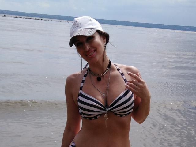 JillianPattie con un bikini a rayas negro y blanco, gorro blanco, collar, sonriendo, con la mano en el pecho, mostrando sus pechos, de pie en agua poco profunda, luciendo linda, día soleado, el agua reflejándose en su piel..