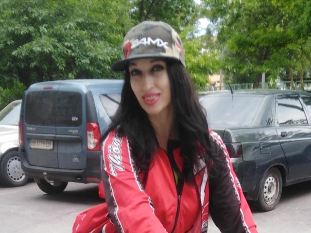 Jillian Pattie, con lunghi capelli neri, indossa un berretto mimetico e una giacca rossa con strisce bianche, sorride con il rossetto rosa, ed è in piedi di fronte a due SUV scuri..