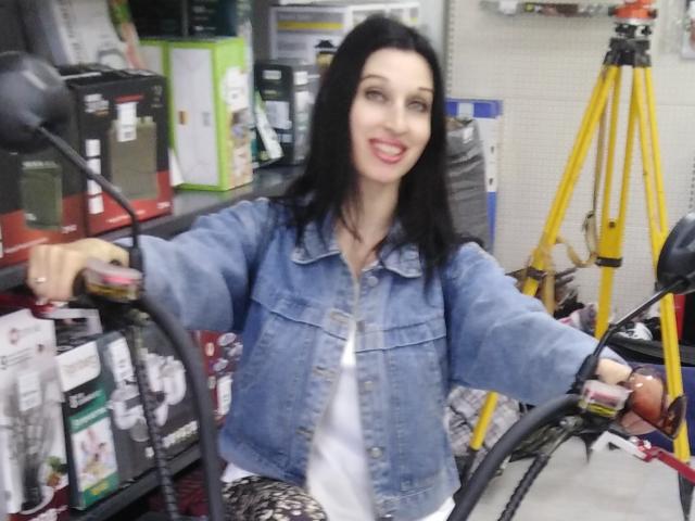 Jillian Pattie mit schwarzen Haaren, einer blauen Jeansjacke, einem weißen Hemd, die Fahrradlenker hält, lächelt, der rechte Arm ist erhoben, sie steht in einem Geschäft, im Hintergrund steht ein gelbes Stativ..