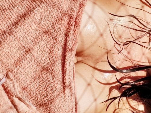 MeahBrunette's wet, shiny tits with glistening skin, covered partially by a pink textured towel, and dark, wet, wavy hair falling over. Sunlight casts diamond-shaped shadows.